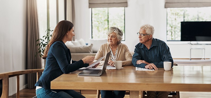 A young woman sitting at a laptop talking to an elderly couple at a kitchen table.